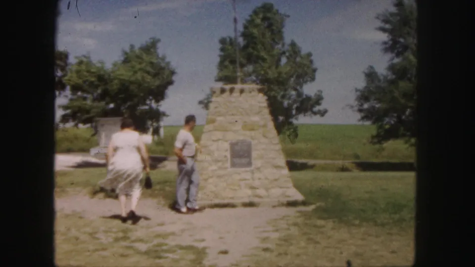 Adult travelers move around a stone roadside monument with trees and open fields behind it in the 1958 Denver-area home movie.
