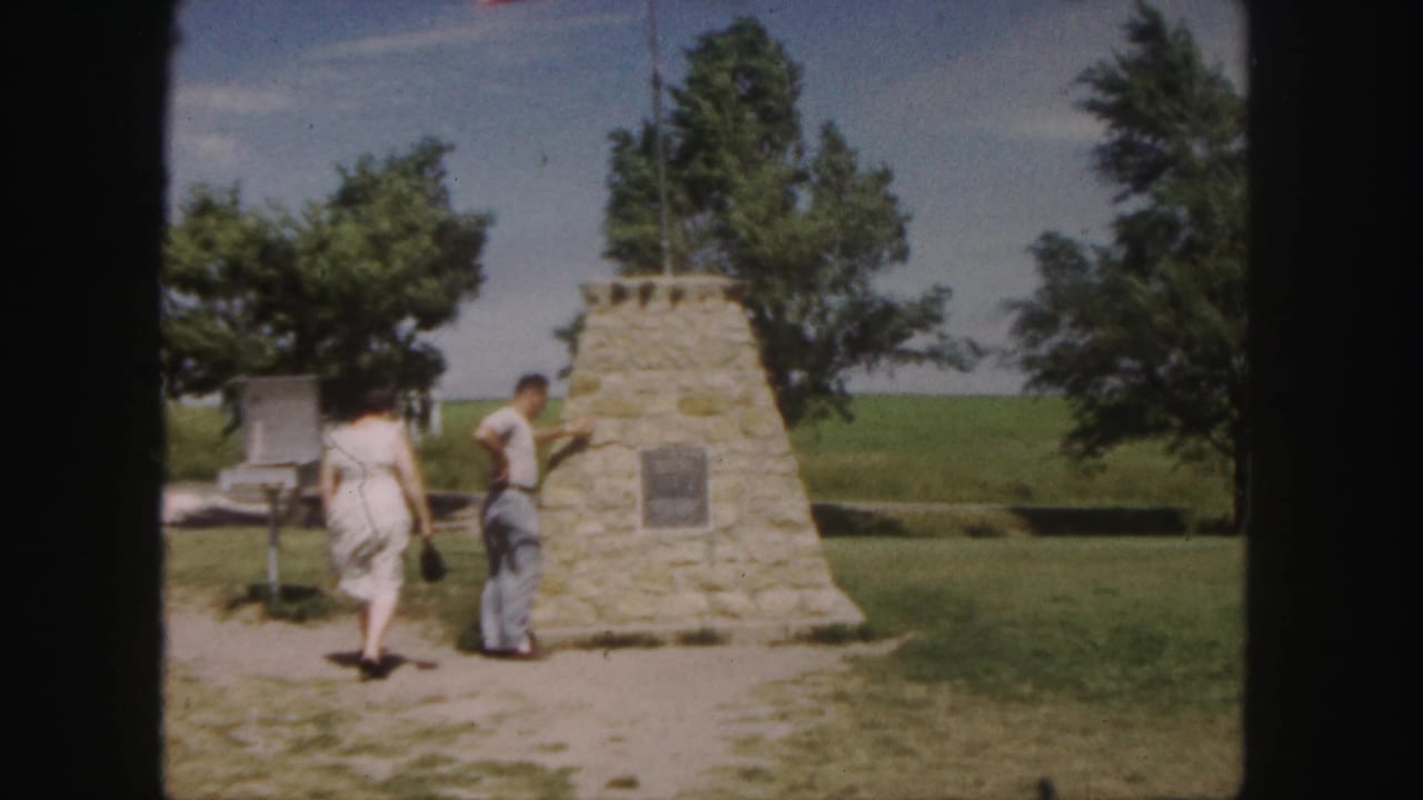 In 1958 Denver home-movie footage, a couple stands by a stone monument with a plaque, flagpole, trees, and open grass.