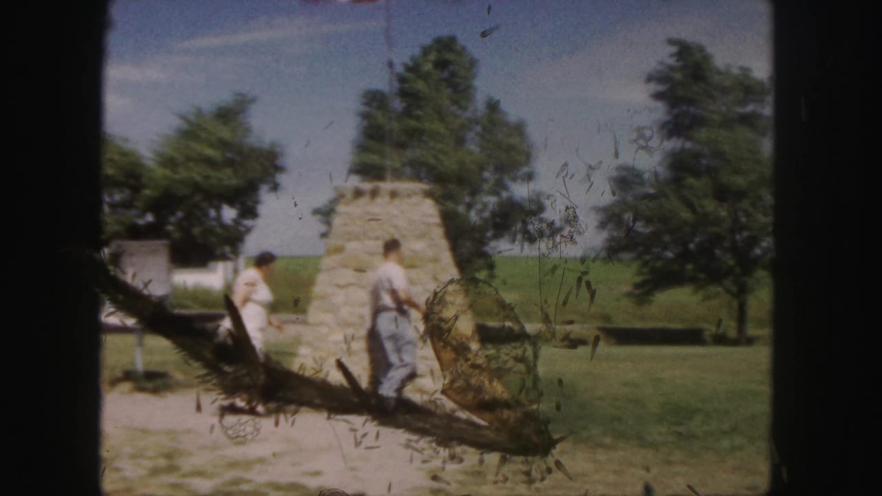 The couple moves around the Denver stone monument while an American flag flies overhead and trees line the background.