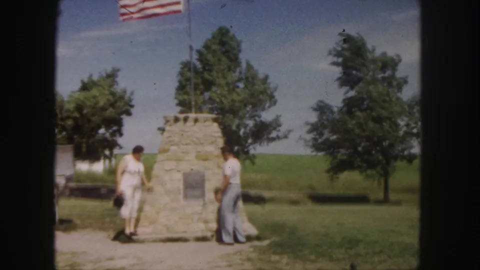 A woman holding a camera and a man stand beside the 1958 Denver monument, with flag, plaque, grass, and trees visible.