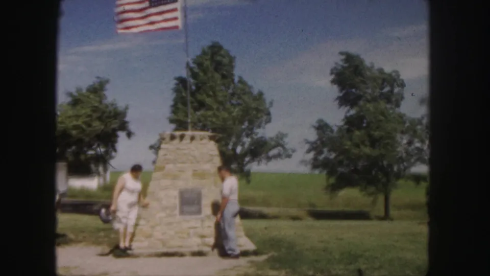 The final frame shows the couple near the Denver stone monument under an American flag, with trees and open grass behind them.
