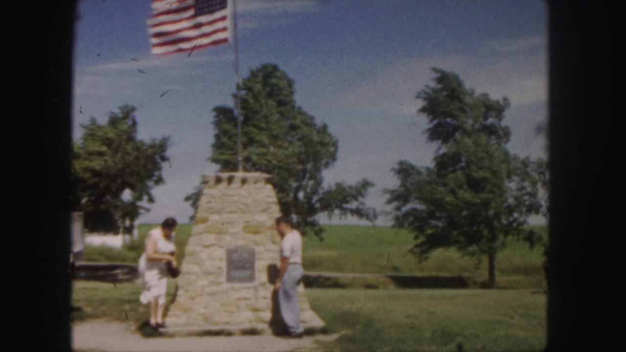 1958 Denver home-movie frame showing two visitors by a stone monument with a plaque, grass, trees, and a U.S. flag overhead.