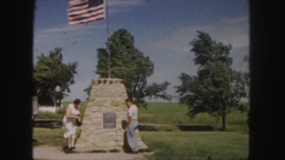 1958 Denver home-movie frame showing two visitors by a stone monument with a plaque, grass, trees, and a U.S. flag overhead.