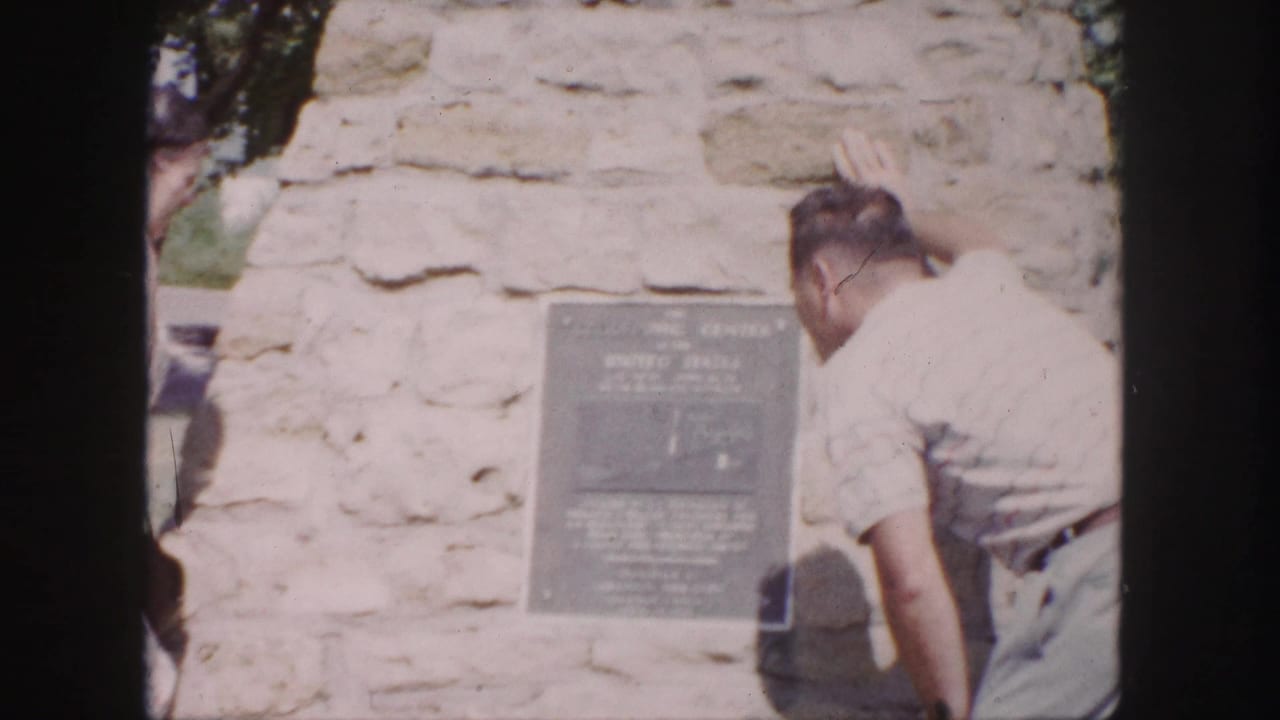 1958 Denver frame with a man reading or examining a plaque fixed to a light stone monument wall.