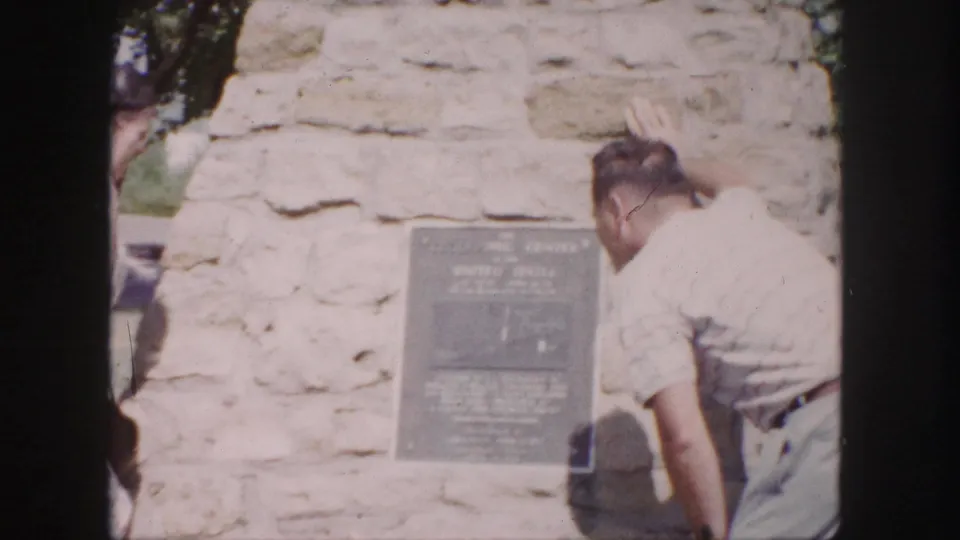1958 Denver frame with a man reading or examining a plaque fixed to a light stone monument wall.