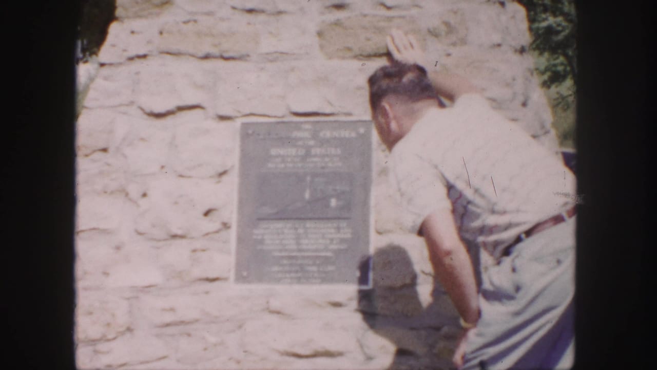 1958 8mm frame of a man leaning close to read a dark plaque on a stone wall, with greenery at the edge of the shot.