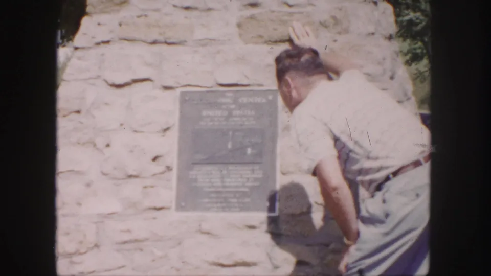 1958 8mm frame of a man leaning close to read a dark plaque on a stone wall, with greenery at the edge of the shot.