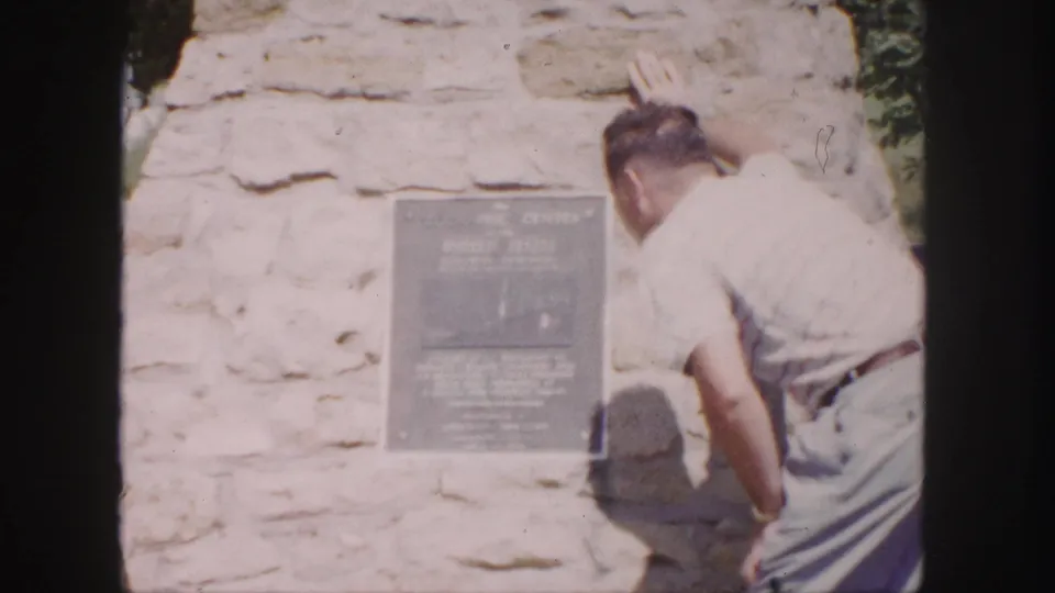 A man in light clothing studies the Geographic Center plaque on a stone wall in a 1958 home-movie frame.