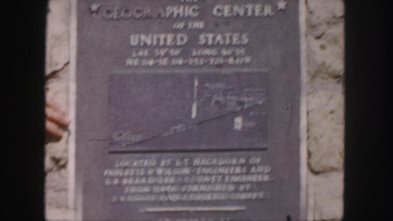 1958 8mm frame of a Geographic Center of the United States plaque mounted on stone, with a partial hand visible at left.