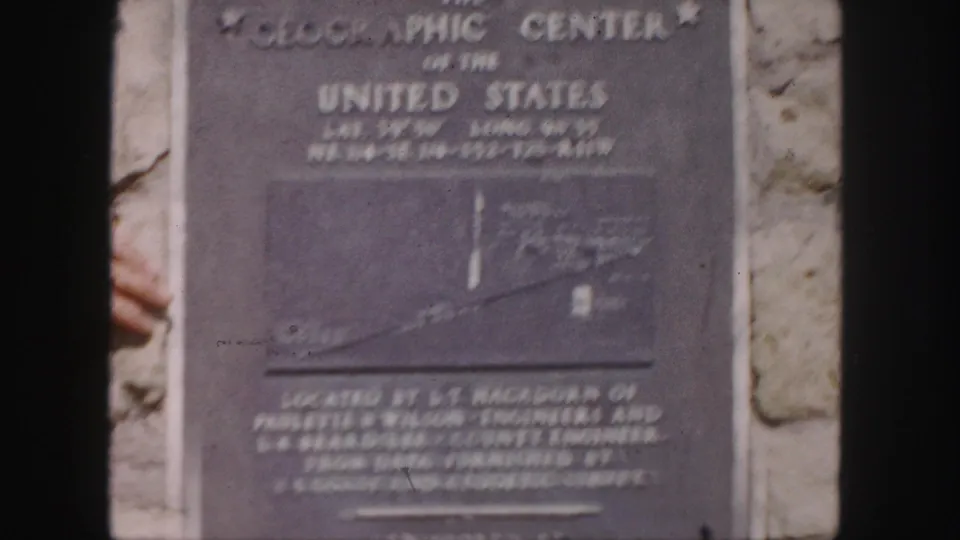 1958 8mm frame of a Geographic Center of the United States plaque mounted on stone, with a partial hand visible at left.