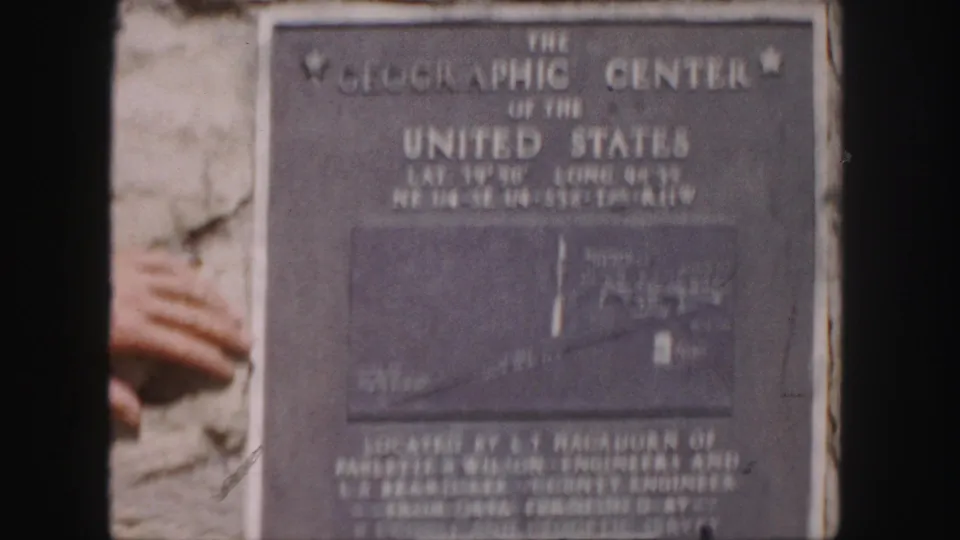 1958 8mm frame of the landmark plaque on stone, slightly soft and faded, with a partial hand along the left border.