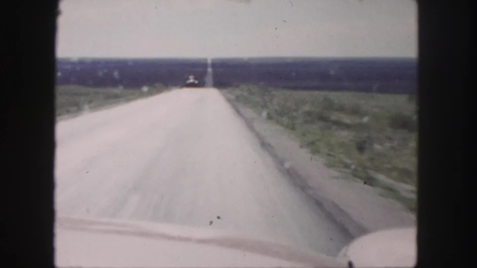 1958 8mm frame looking over a vehicle hood down a straight rural road beneath a bright sky.