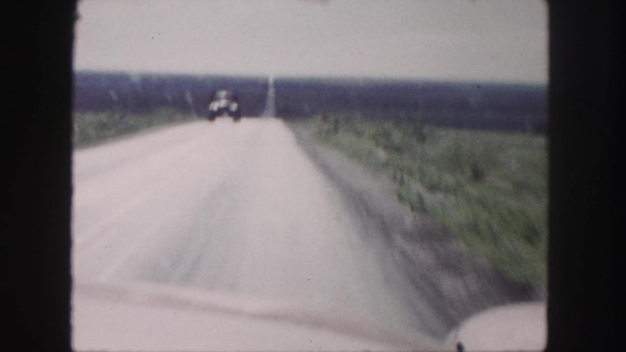 Distant oncoming car appears on a straight road bordered by open fields.