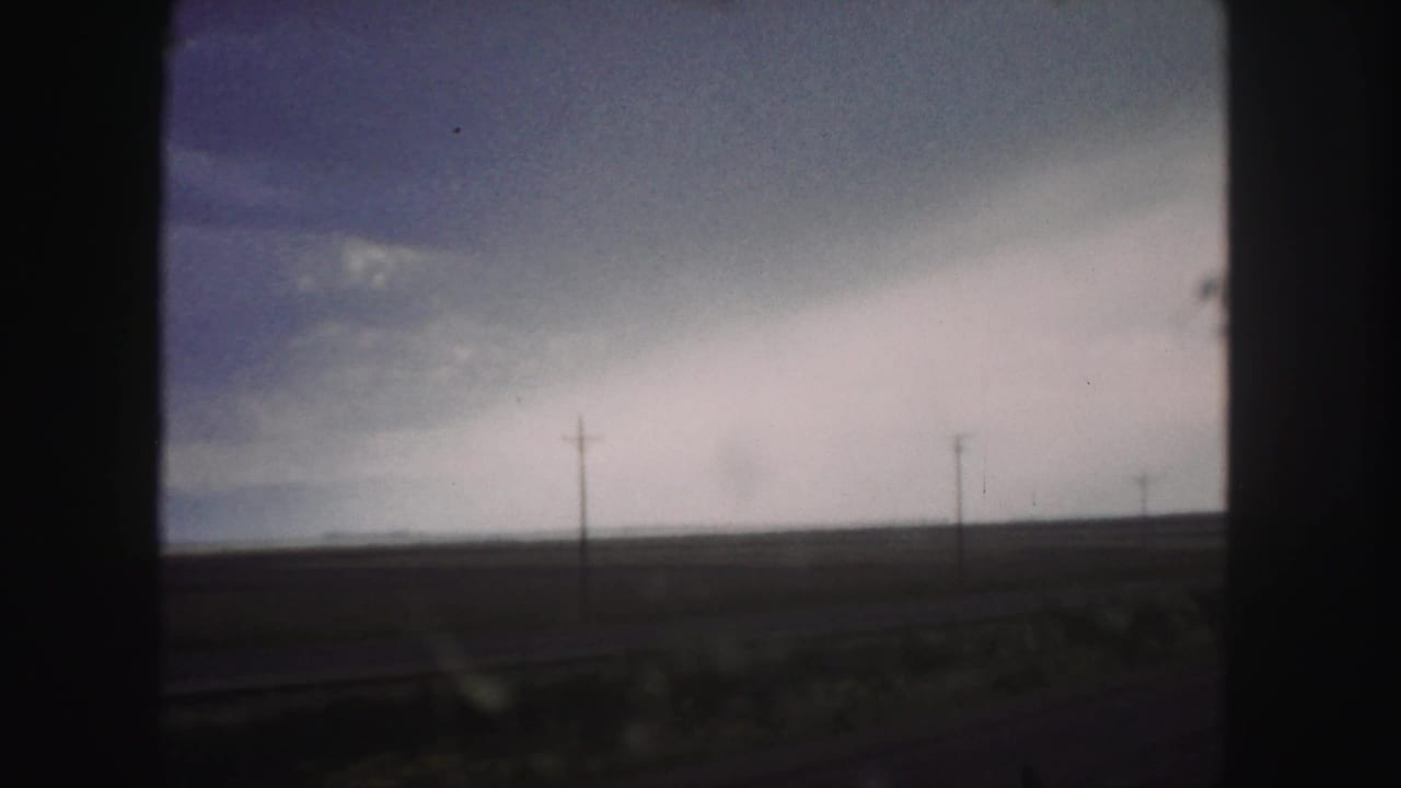 Dark-framed view of open land, utility poles, and a heavy cloud bank on the horizon.