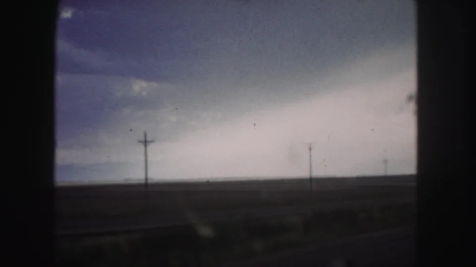 1958 Denver-area moving-vehicle view of a flat highway-side landscape, utility poles, and layered blue-gray clouds.