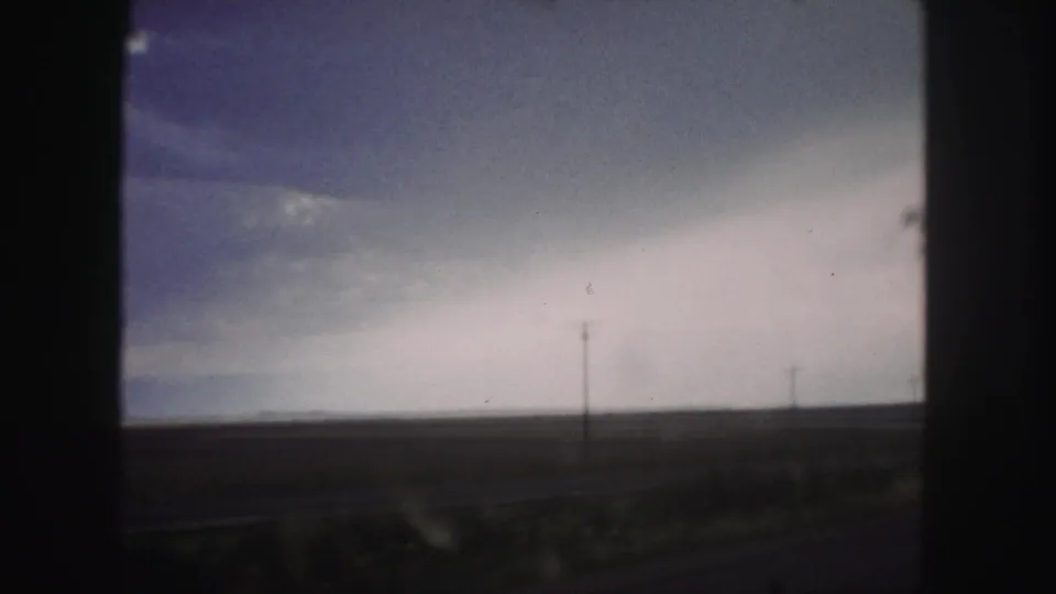 1958 Denver-area 8mm road-travel frame with utility poles, open land, and a cloudy sky viewed through a vehicle window.