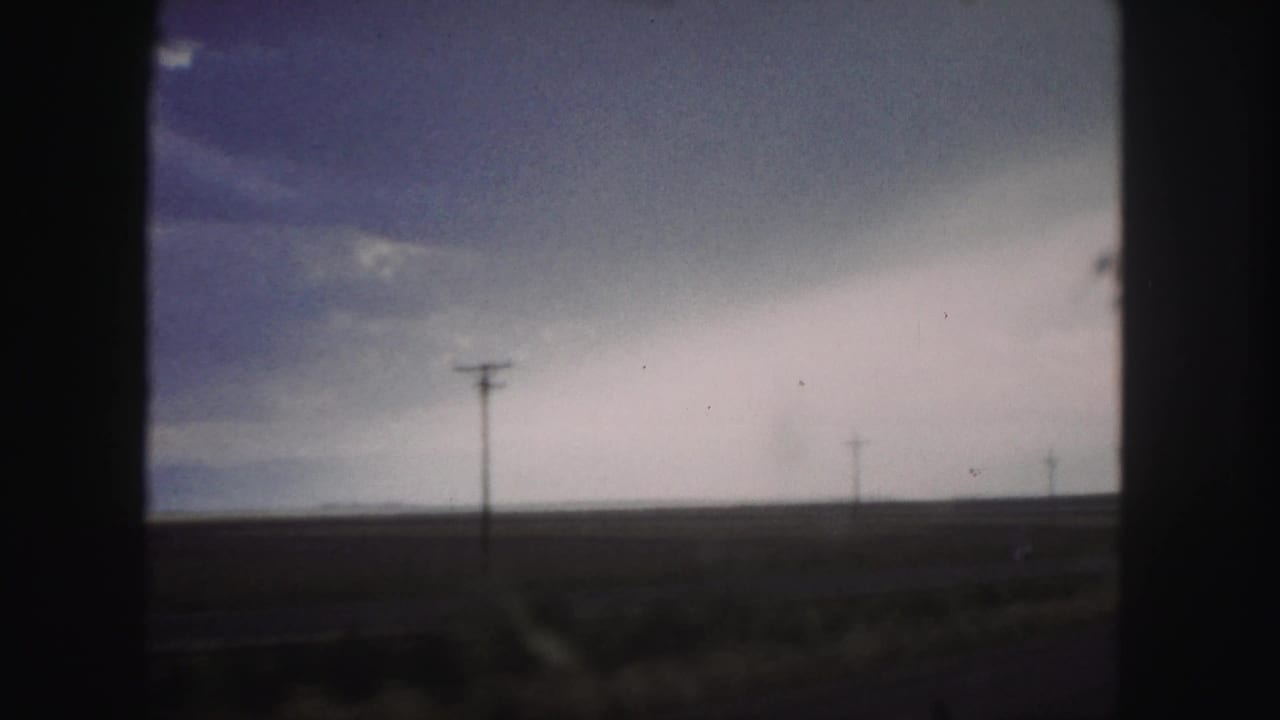 Denver-area 1958 roadside landscape with open fields, distant utility poles, and a muted cloudy sky filmed from a moving vehicle.