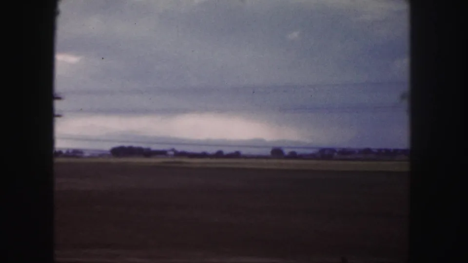 1958 Denver-area passing view of flat fields with a distant tree line or buildings under layered gray clouds.