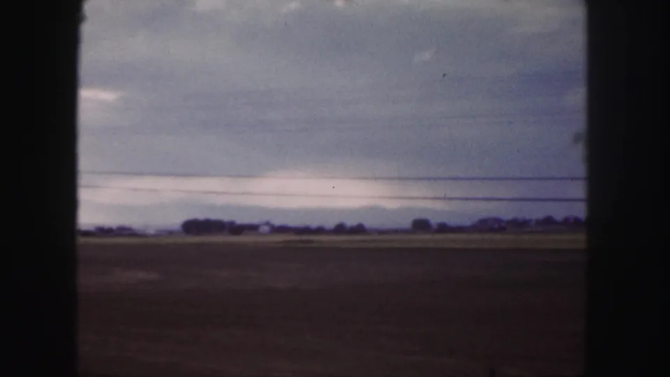 1958 Colorado home-movie frame showing flat plains, distant low buildings, and power lines beneath a clouded sky.