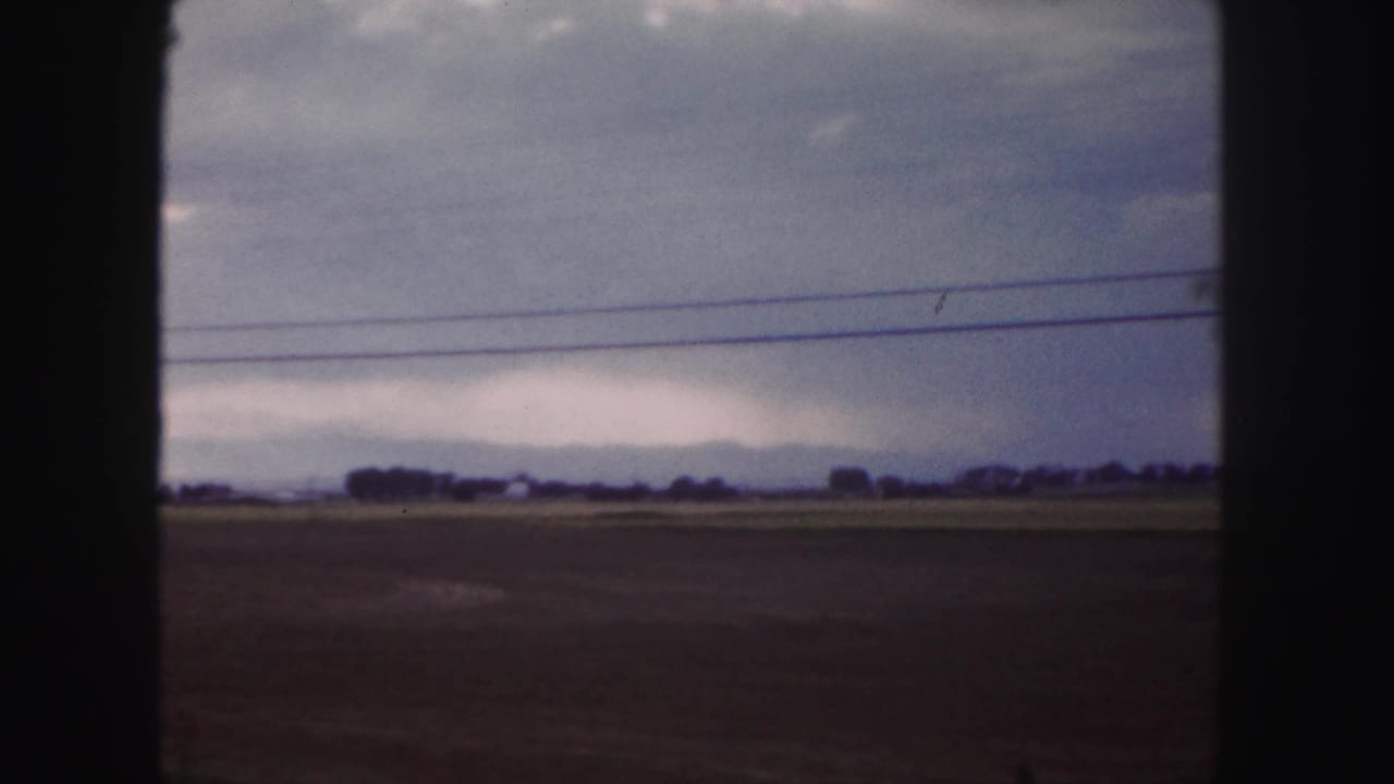 Moving 8mm view near Denver showing open fields, a distant horizon, and roadside power lines under heavy clouds.