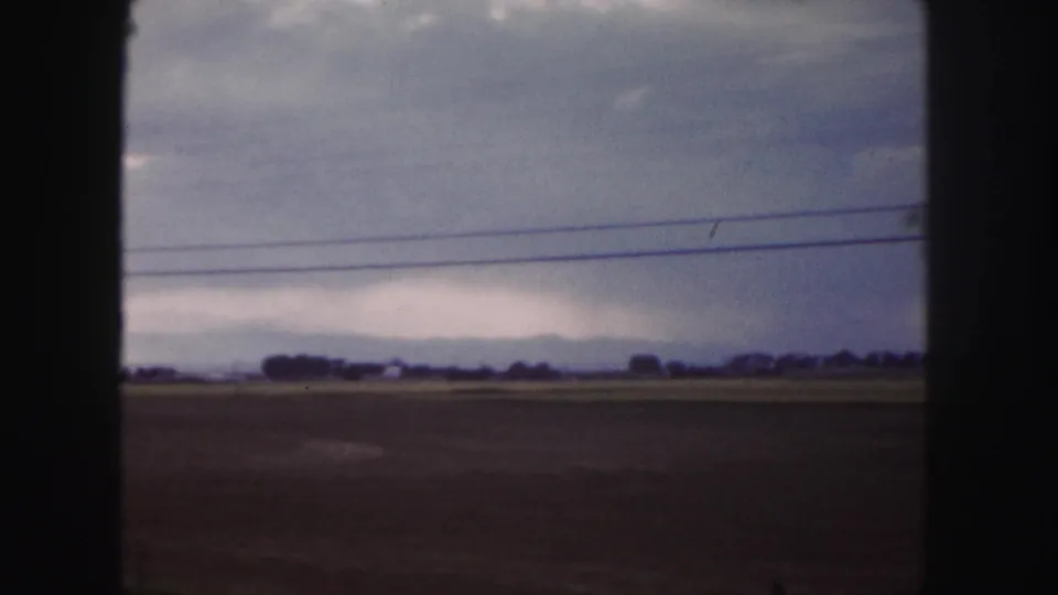 Moving 8mm view near Denver showing open fields, a distant horizon, and roadside power lines under heavy clouds.