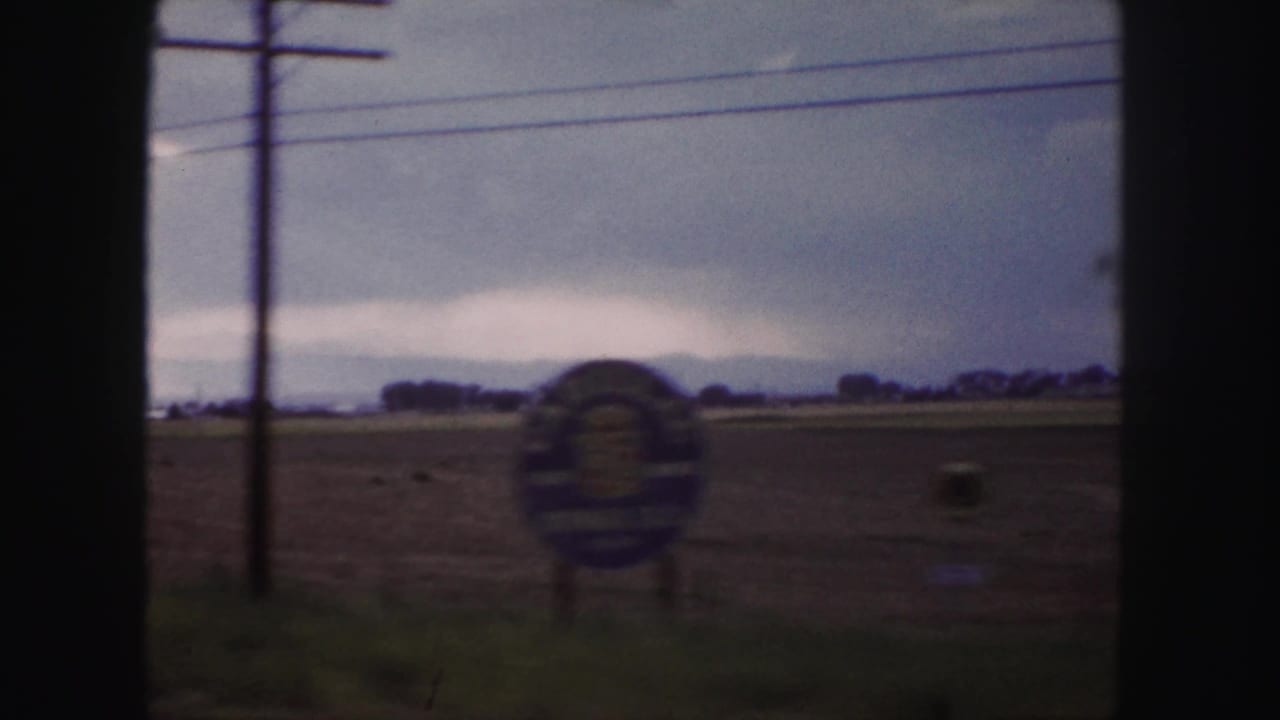 1958 Denver-area home-movie frame showing a soft roadside view with poles, open ground and a partially legible roadside sign.