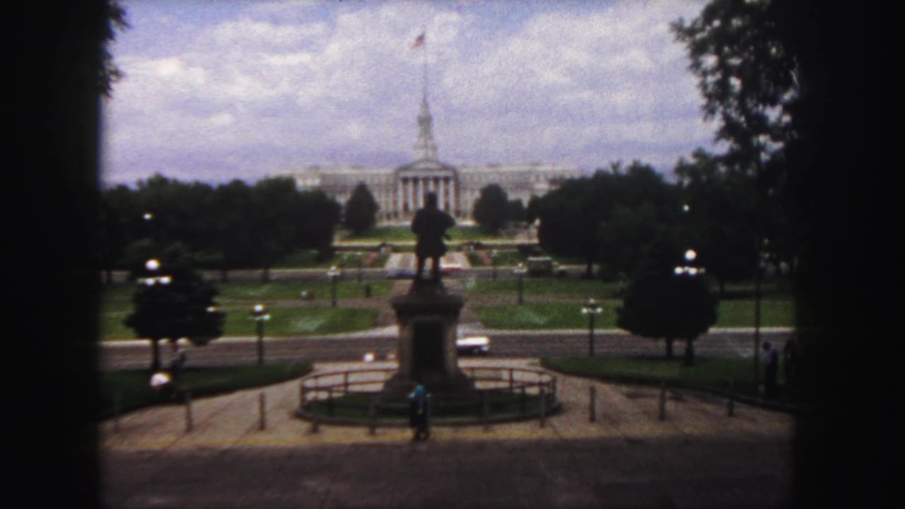 Statue in a plaza with lawns, trees, cars and a classical building beyond.
