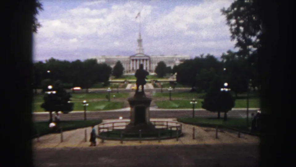 Denver, 1958: a shaded home-movie view of a plaza statue, surrounding trees, cars and a distant spired civic building.