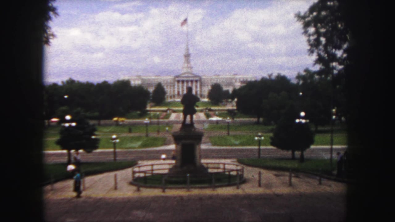 Capitol building beyond Civic Center Park with statue, trees, paths, lampposts, and small pedestrians.