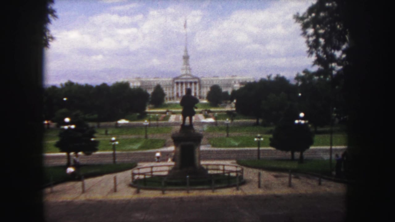 1958 Denver view of a distant classical civic building, silhouetted statue, lawns, trees, roadway, and cloudy sky on soft 8mm film.