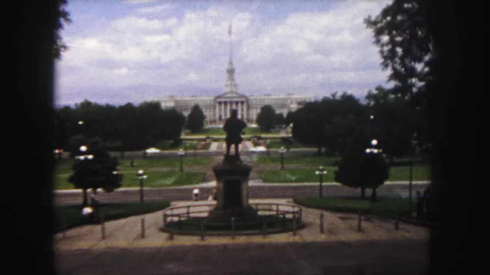 1958 Denver view of a distant classical civic building, silhouetted statue, lawns, trees, roadway, and cloudy sky on soft 8mm film.