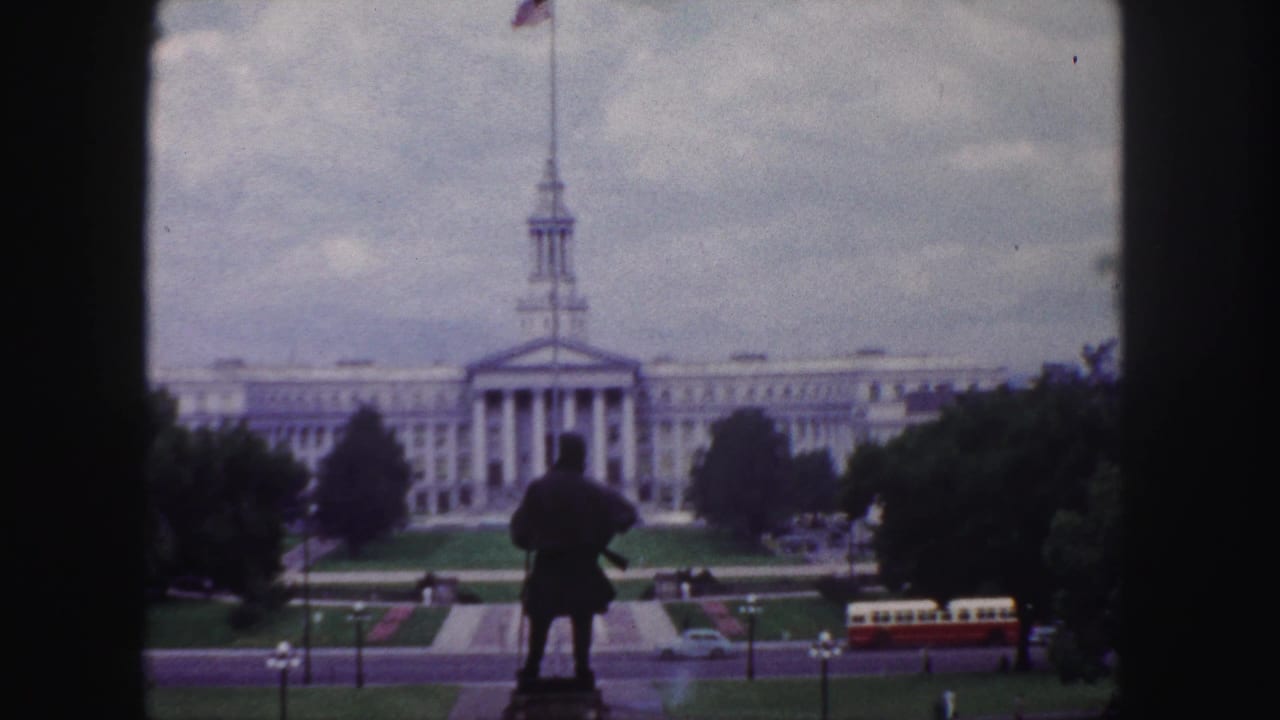 Statue silhouette before a large classical building as a red-and-cream bus passes on the roadway.