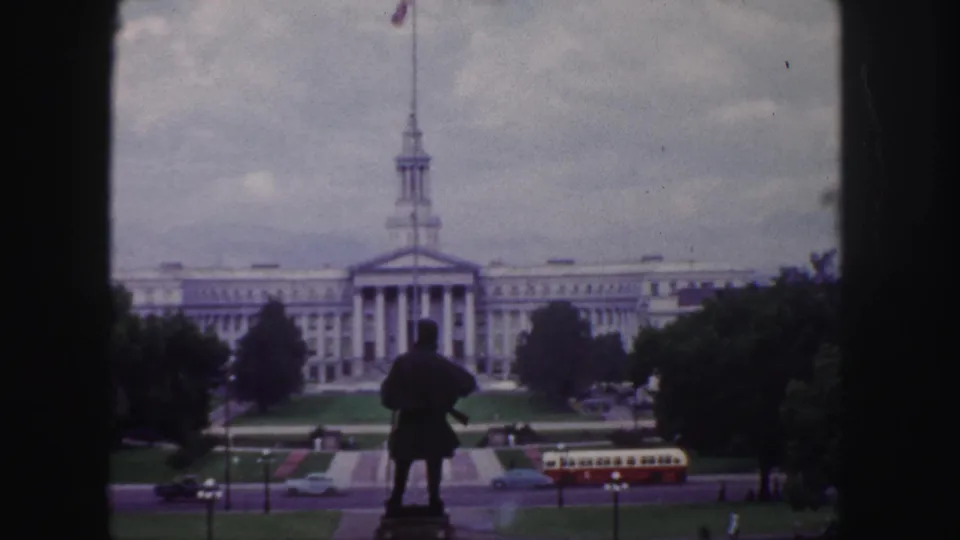 1958 Denver home-movie view across a statue and landscaped grounds toward a broad classical building with cars and a bus below.