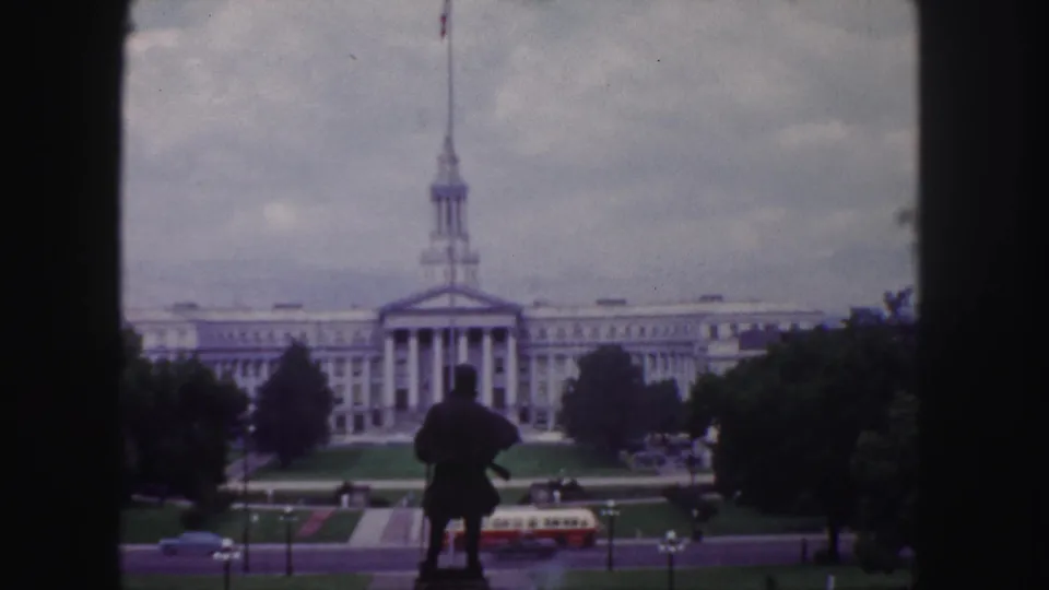 Soft 8mm Denver frame showing a silhouetted statue, classical public building, landscaped grounds, trees, roadway traffic, and cloudy sky.