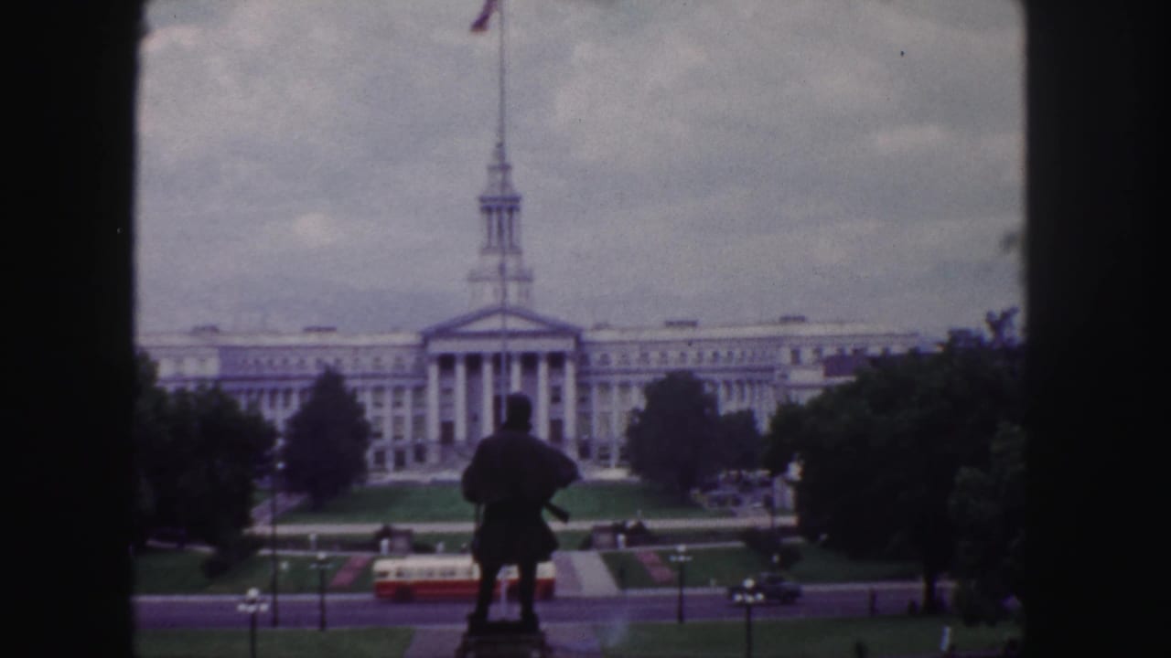 1958 Denver 8mm frame showing a spired civic-style building beyond a statue, with lawns, road traffic, and a red-and-white bus below.