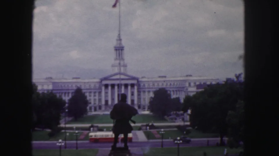 1958 Denver 8mm frame showing a spired civic-style building beyond a statue, with lawns, road traffic, and a red-and-white bus below.