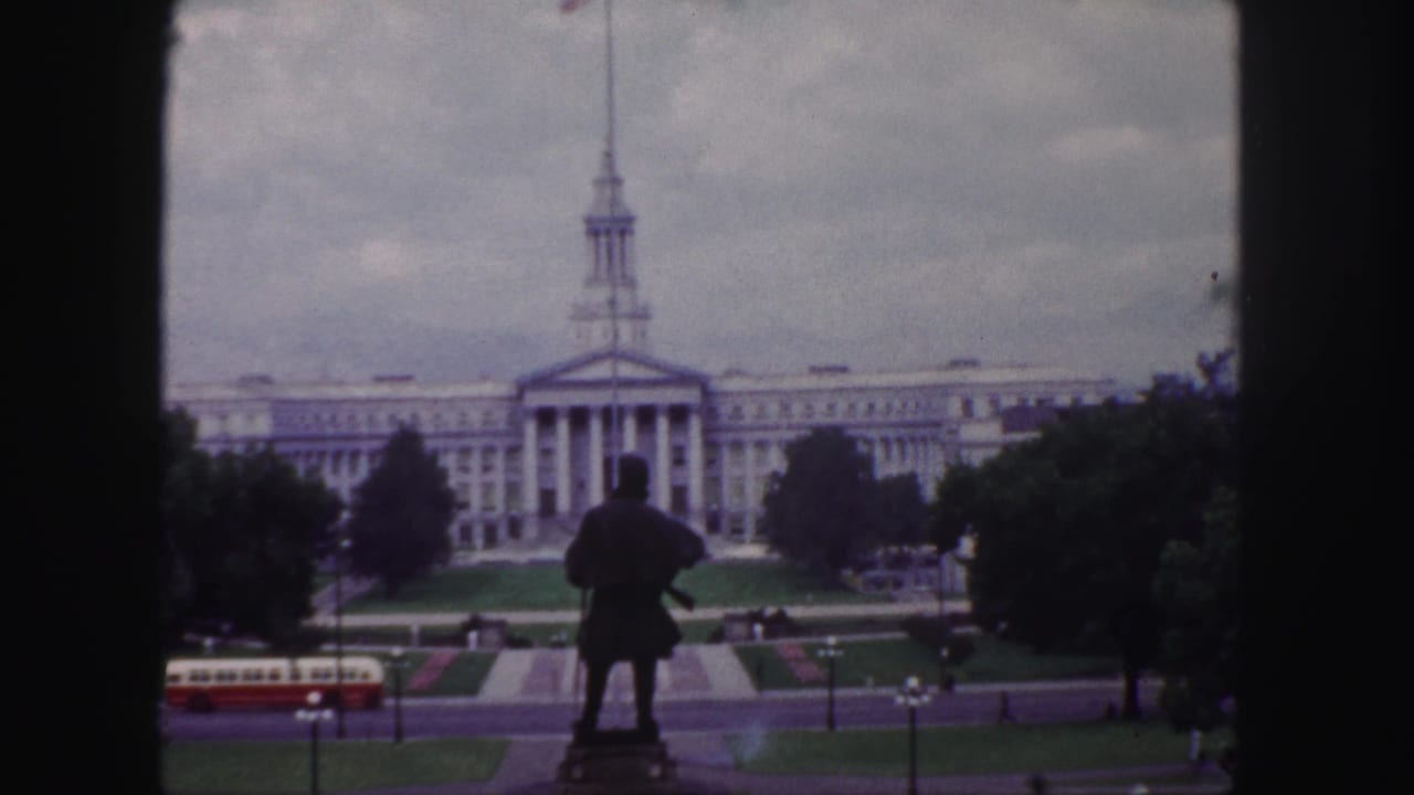 1958 Denver view through dark frame edges toward a spired civic-style building, with a statue, formal grounds, trees, and bus traffic below.
