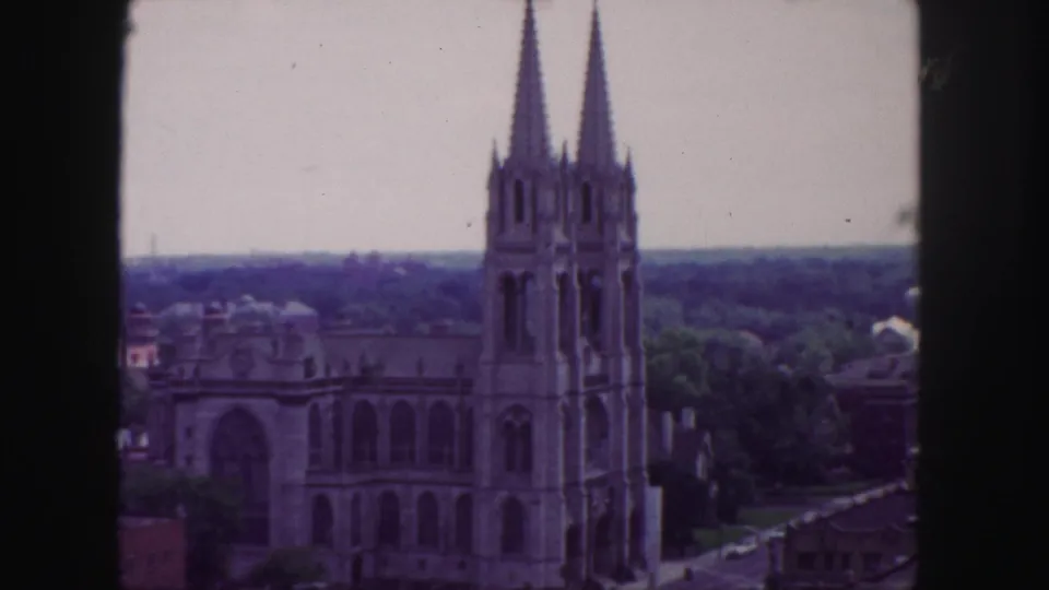 1958 Denver 8mm frame showing a twin-spired church-like building from an elevated viewpoint, with trees, rooftops, and a distant skyline.