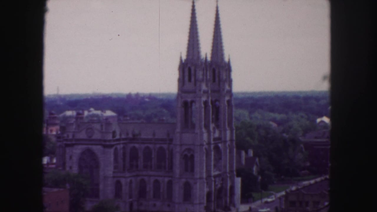 1958 Denver 8mm frame showing a Gothic cathedral with twin spires, trees, rooftops, and a hazy skyline from an elevated viewpoint.