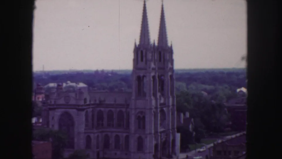 1958 Denver 8mm frame showing a Gothic cathedral with twin spires, trees, rooftops, and a hazy skyline from an elevated viewpoint.