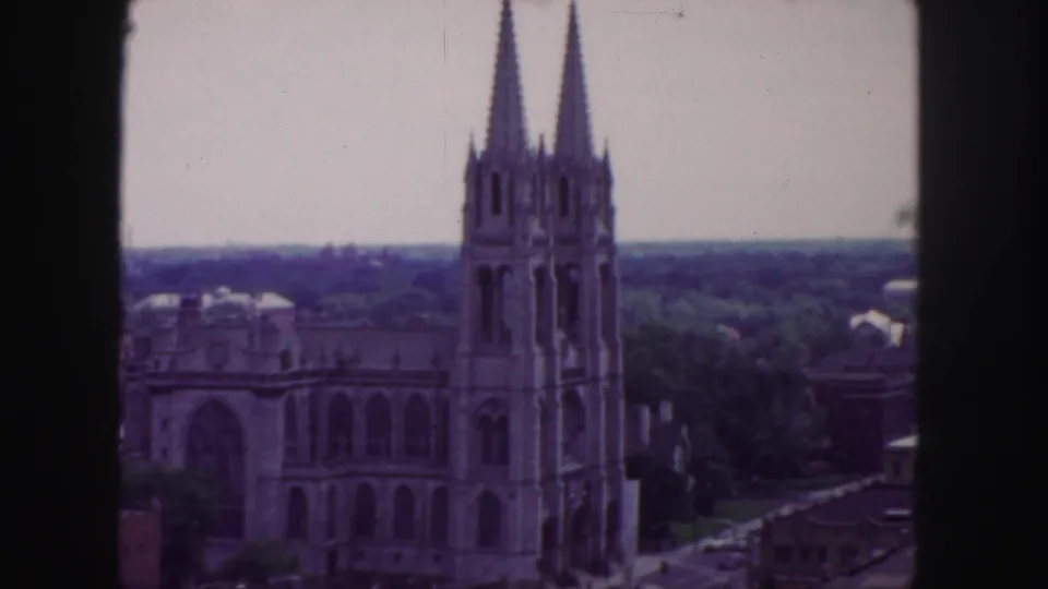 Denver 1958 8mm still with soft color showing the cathedral's twin spires, arched front, leafy grounds, and surrounding rooftops.