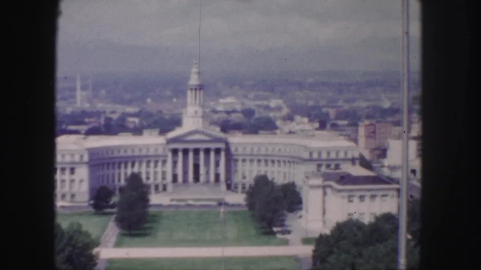 1958 Denver frame showing a broad civic building with a portico, formal lawn, paths, trees, and a hazy city background.