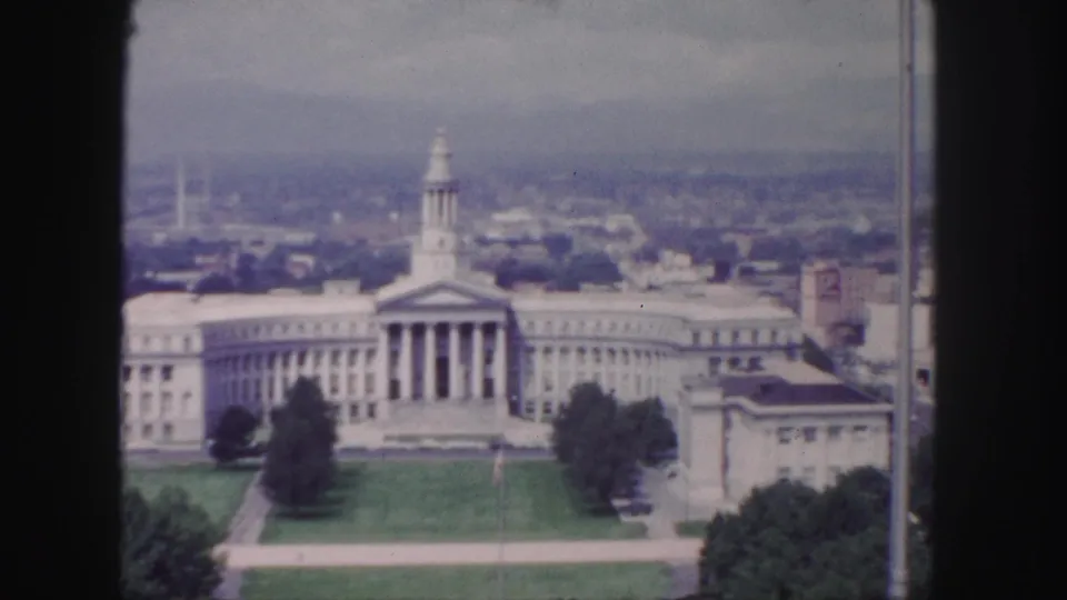 1958 Denver home-movie frame of a columned civic building with central tower, broad lawn, trees, and dark vertical film borders.