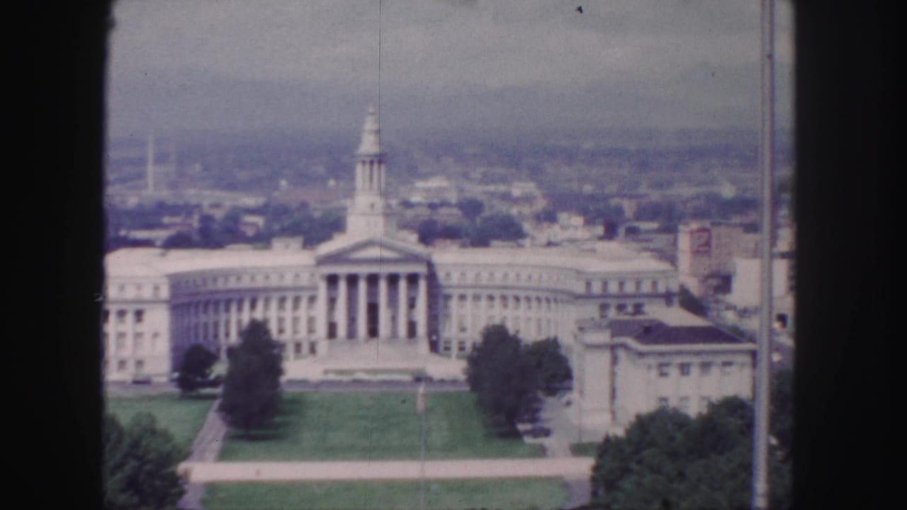 Elevated view of a large columned building with wings, lawn, trees, and hazy city background.
