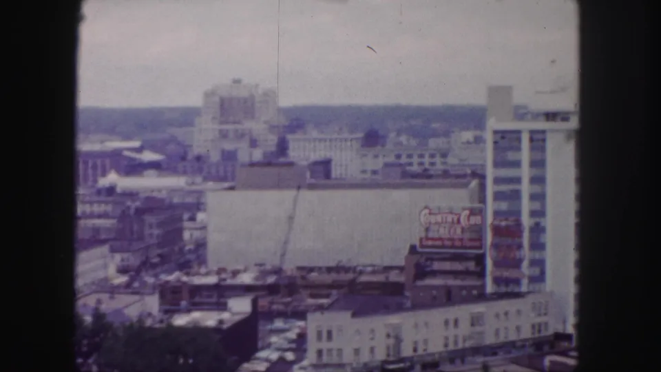 1958 Denver home-movie frame looking across downtown rooftops, with a readable commercial sign, taller buildings, and hazy distance.