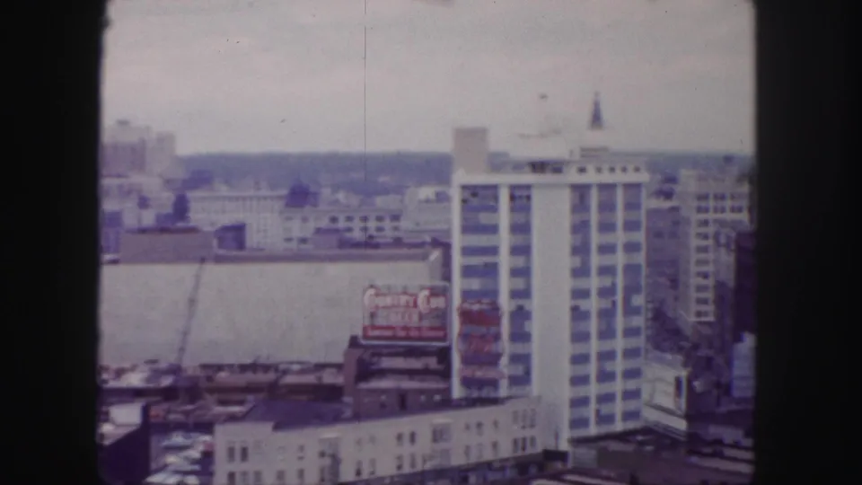 Panoramic 1958 Denver view with the camera shifted across construction, a beer billboard, rooftops, and a tall modern high-rise.