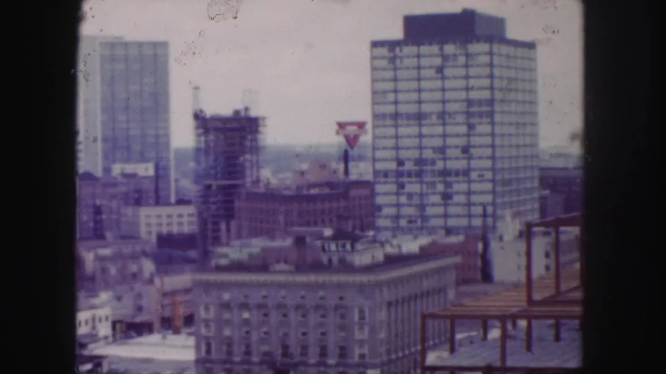 Later 1958 Denver skyline frame with a gridded high-rise, construction-like structure, rooftop signage, and older downtown buildings.