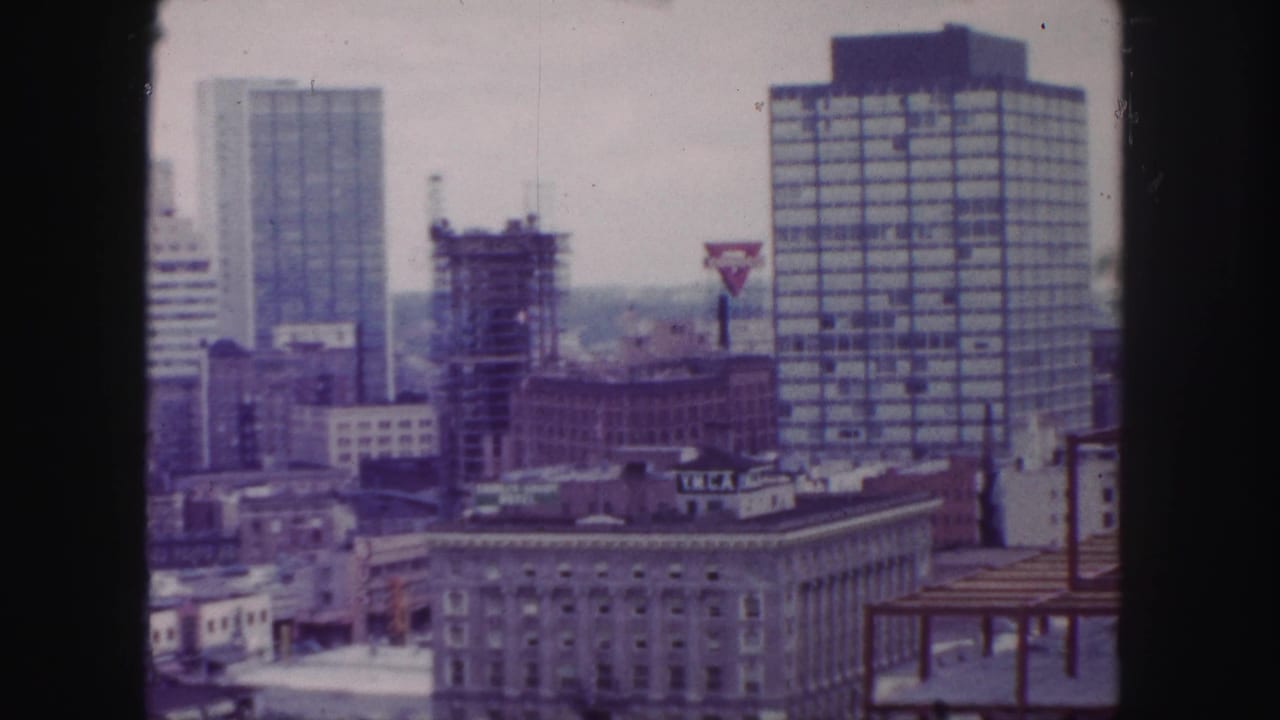 City skyline with rectangular towers, construction scaffolding, rooftop signage, and dark film borders.