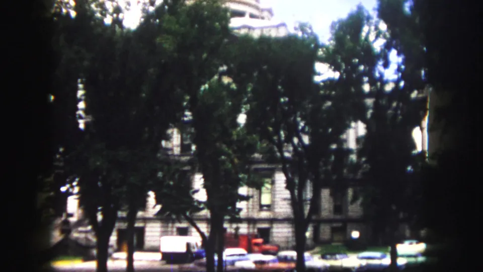 Late-1950s Denver street scene with parked cars, dense tree canopy, and downtown building fronts in soft 8mm focus.