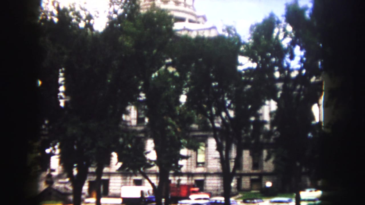 Denver Colorado State Capitol appears behind dark tree trunks and foliage in 1958 8mm footage, with small cars near the base.